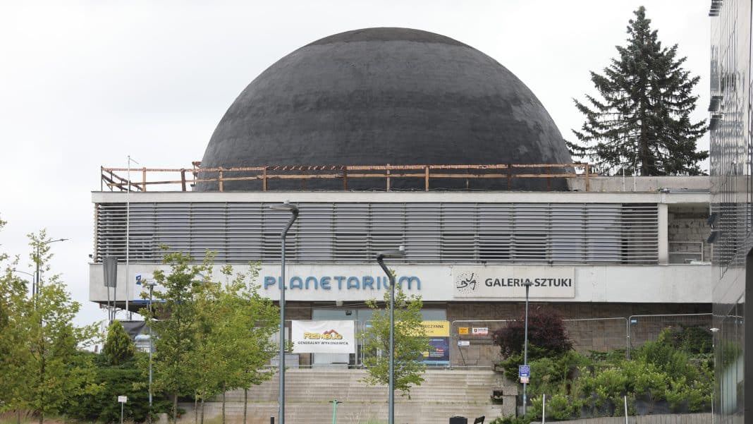 Widok na budynek planetarium z charakterystycznym kopułowym dachem, otoczony drzewami i miejską infrastrukturą.
