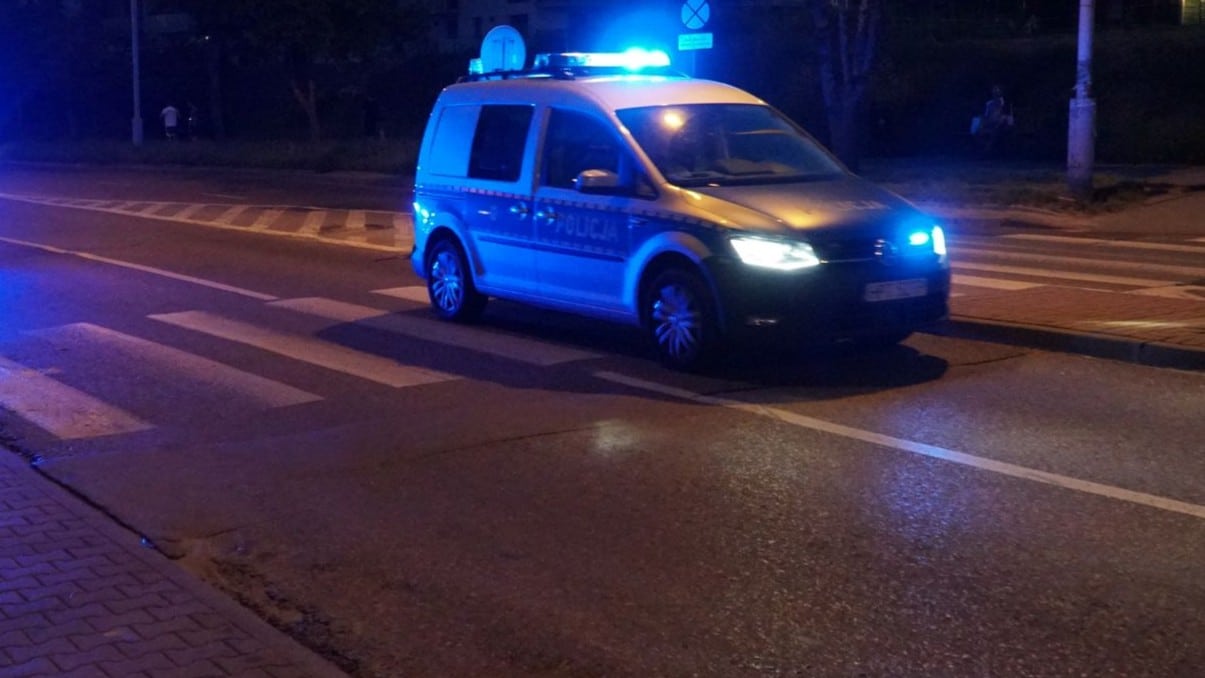 Samochód policyjny z włączonymi światłami na ulicy w nocy, na pasach dla pieszych.