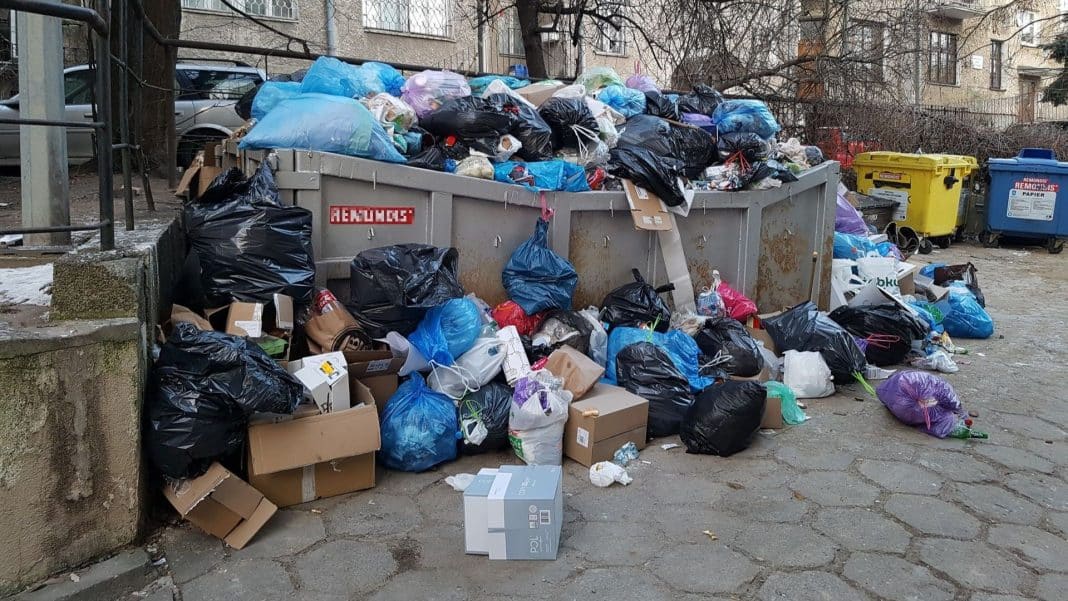 Wielki kontener na śmieci pełen odpadów, wokół porozrzucane worki i kartony, na tle budynków mieszkalnych.