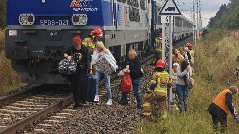 Grupa pasażerów opuszcza pociąg na torze kolejowym, w pobliżu służb ratunkowych i pracowników technicznych.
