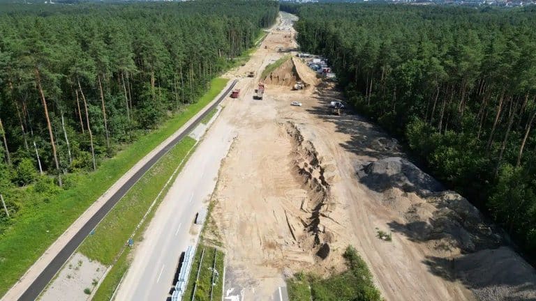 Zdjęcie przedstawia odcinek drogi w trakcie budowy, z wykopami i ciężkim sprzętem, otoczony lasem.