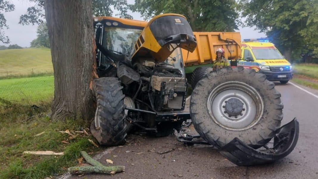 Ciągnik rolniczy uderzył w drzewo na drodze wojewódzkiej, zablokowana jezdnia, w tle pojazd ratunkowy.