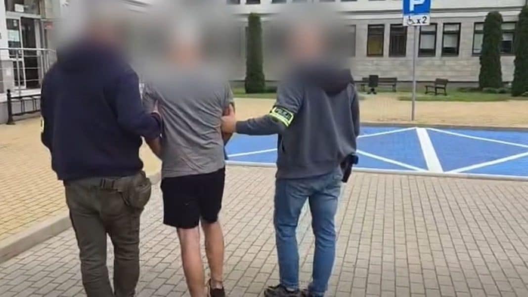 Trzech mężczyzn prowadzi zatrzymanego, jeden z nich ma na ramieniu odznakę policyjną, na tle budynku i parkingu.