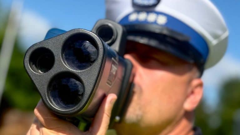 Policjant trzyma radar, na głowie ma hełm, w tle niebo i drzewa.