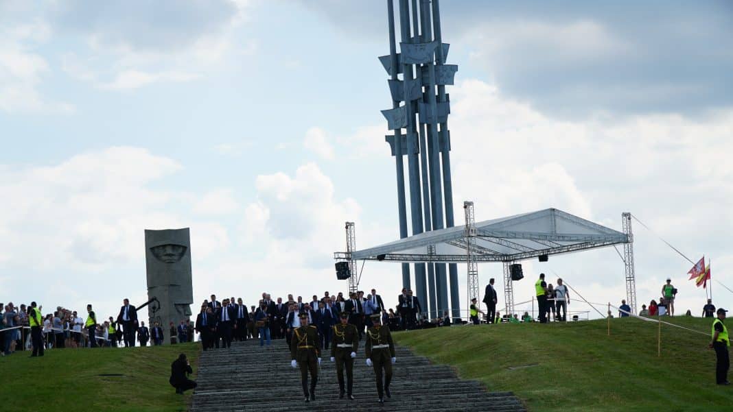 Widok na Pomnik Grunwaldu z grupą ludzi i żołnierzami na schodach, w tle chmury na niebie.