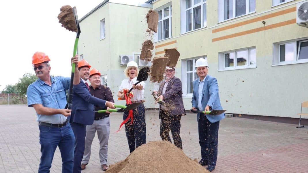 Grupa ludzi w kaskach budowlanych podczas ceremonii rozpoczęcia budowy nowego skrzydła w ośrodku edukacyjnym, z wykopem i łopatami.