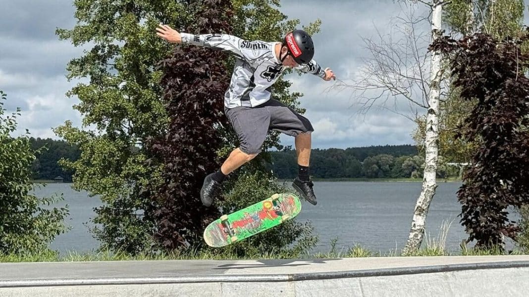 Młody skateboardzista wykonuje trik na skateparku z jeziorem i drzewami w tle.