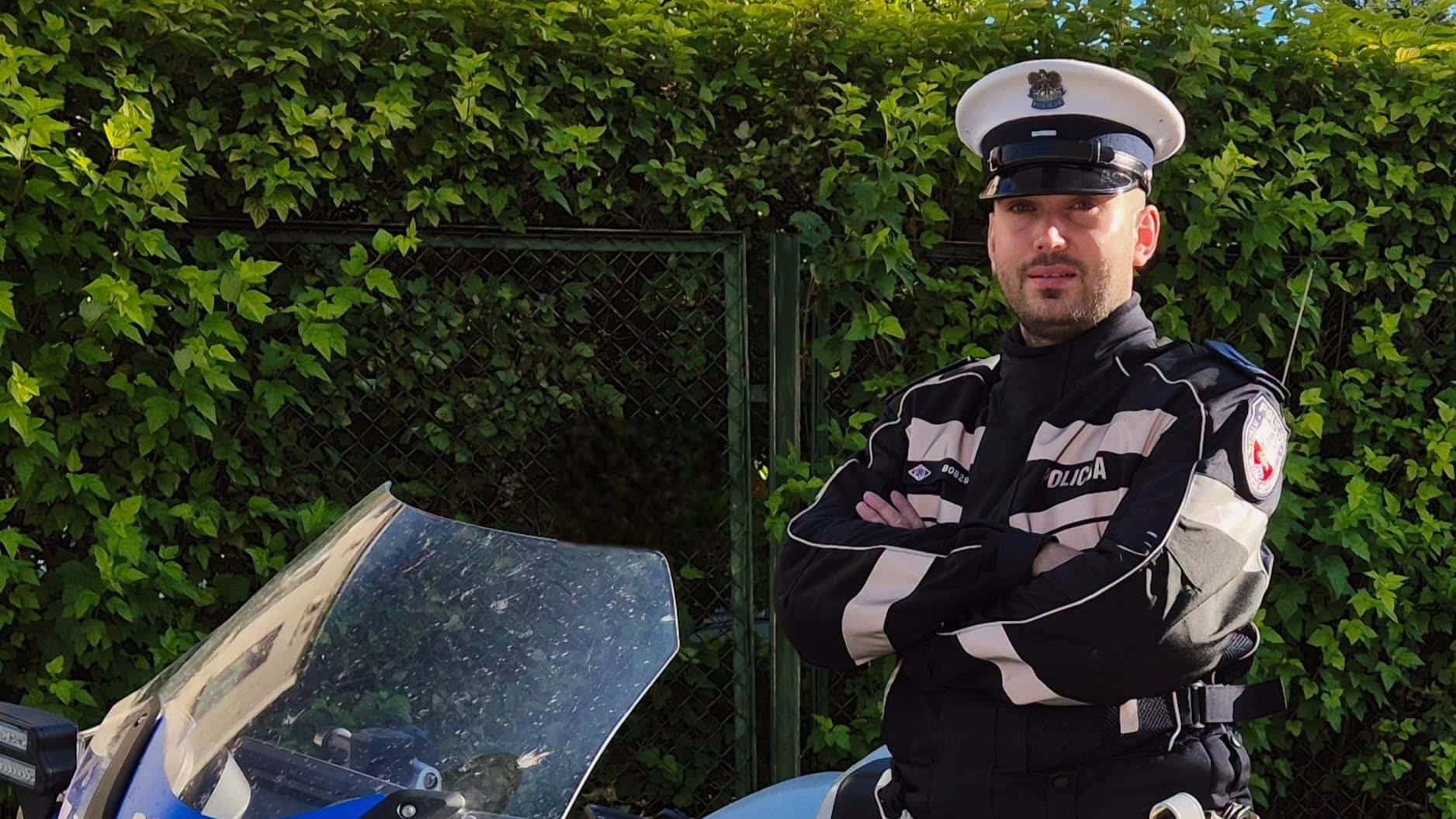 Policjant w mundurze stoi obok motoru, za nim zielona roślinność i metalowa brama.