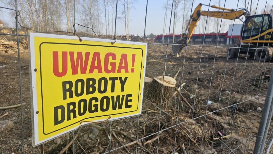 Zdjęcie placu budowy z ogrodzeniem i tablicą ostrzegawczą, w tle koparka i wykopane ziemie.