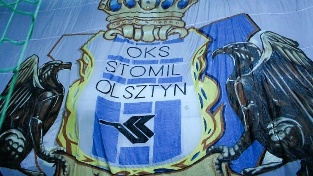 Na zdjęciu emblemat klubu piłkarskiego OKS Stomil Olsztyn z herbem i motywami orłów po bokach.