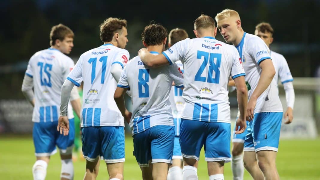 Grupa piłkarzy Stomilu Olsztyn w białych koszulkach i niebieskich spodenkach na boisku, podczas meczu.