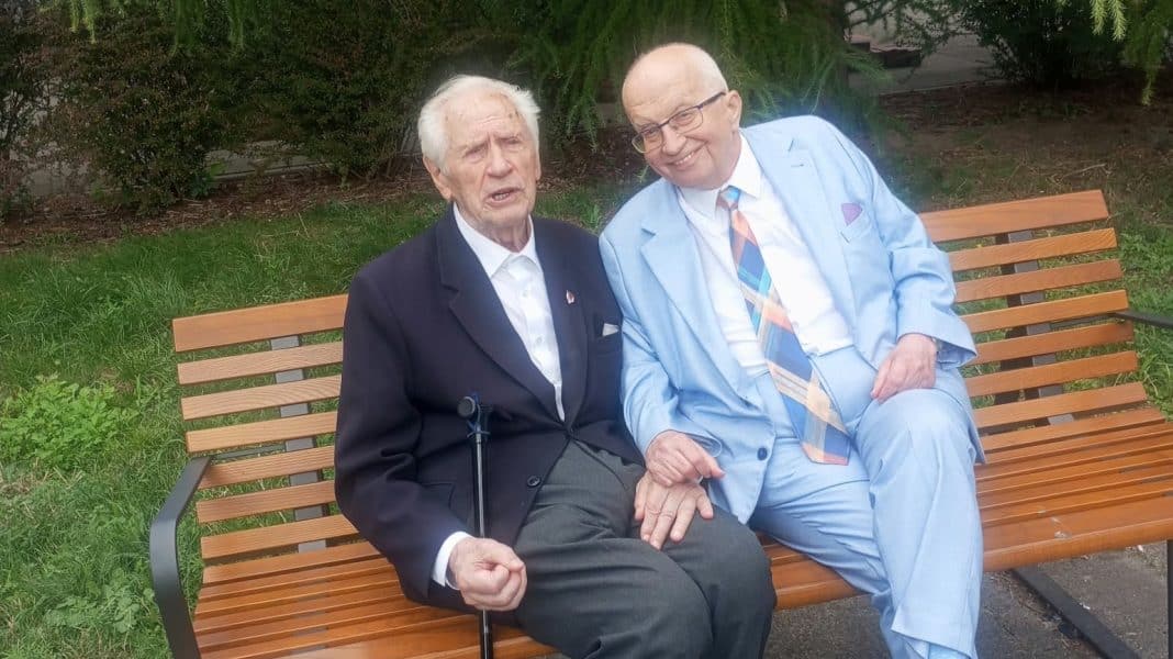 Dwoje starszych mężczyzn siedzi na ławce w ogrodzie, trzymając się za ręce, jeden z nich korzysta z kul.