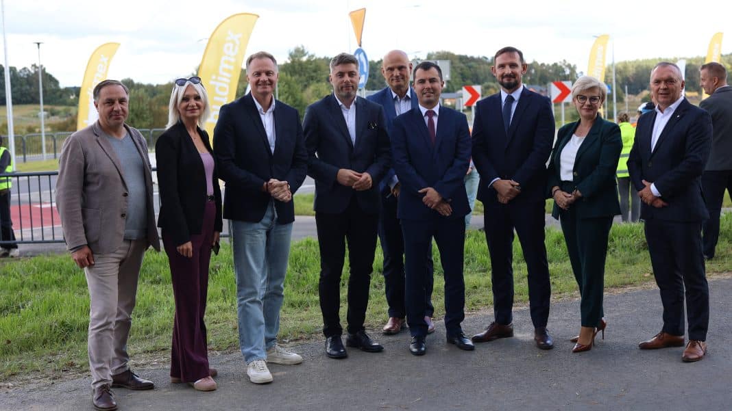 Grupa ludzi na otwarciu nowej drogi na Warmii i Mazurach, w tle znaki drogowe i flagi.