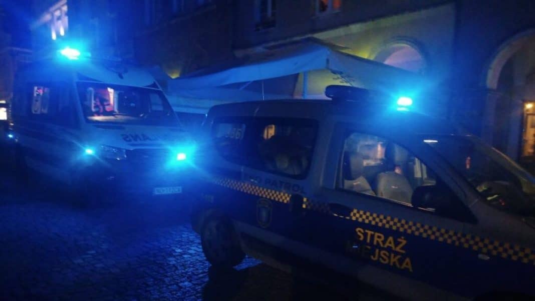 Na zdjęciu widać radiowozy straży miejskiej z włączonymi niebieskimi światłami nocą na ulicy.