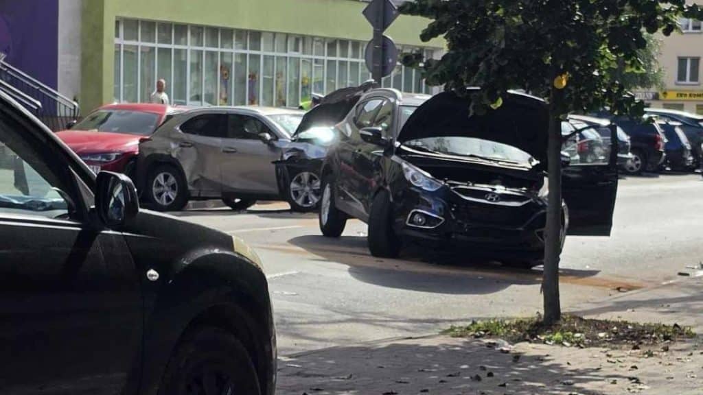Zdjęcie przedstawia rozbite trzy samochody na parkingu, w tym czarny samochód z otwartym maską.