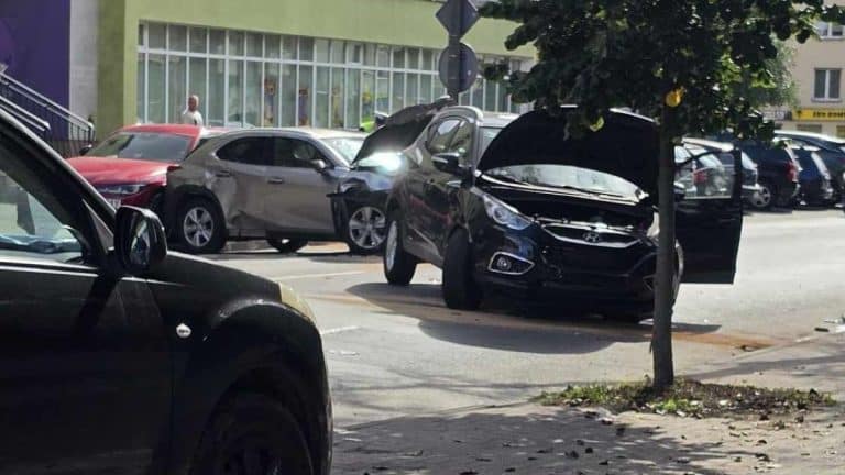 Zdjęcie przedstawia rozbite trzy samochody na parkingu, w tym czarny samochód z otwartym maską.