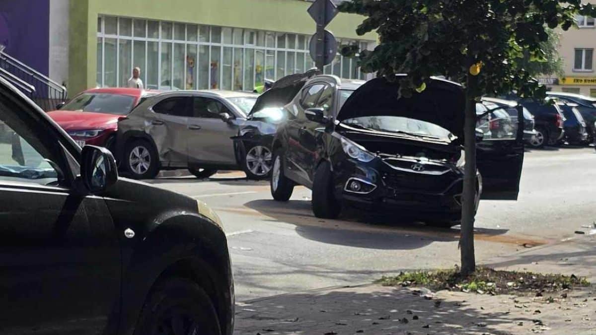 Zdjęcie przedstawia rozbite trzy samochody na parkingu, w tym czarny samochód z otwartym maską.