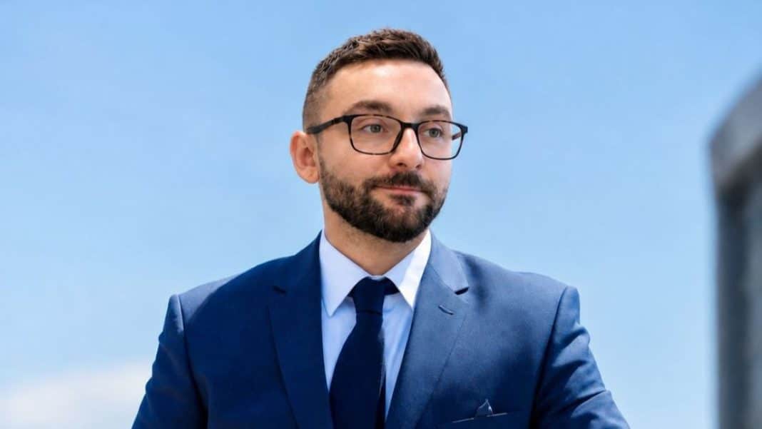 Mężczyzna w garniturze i okularach na tle nieba, patrzący w bok.
