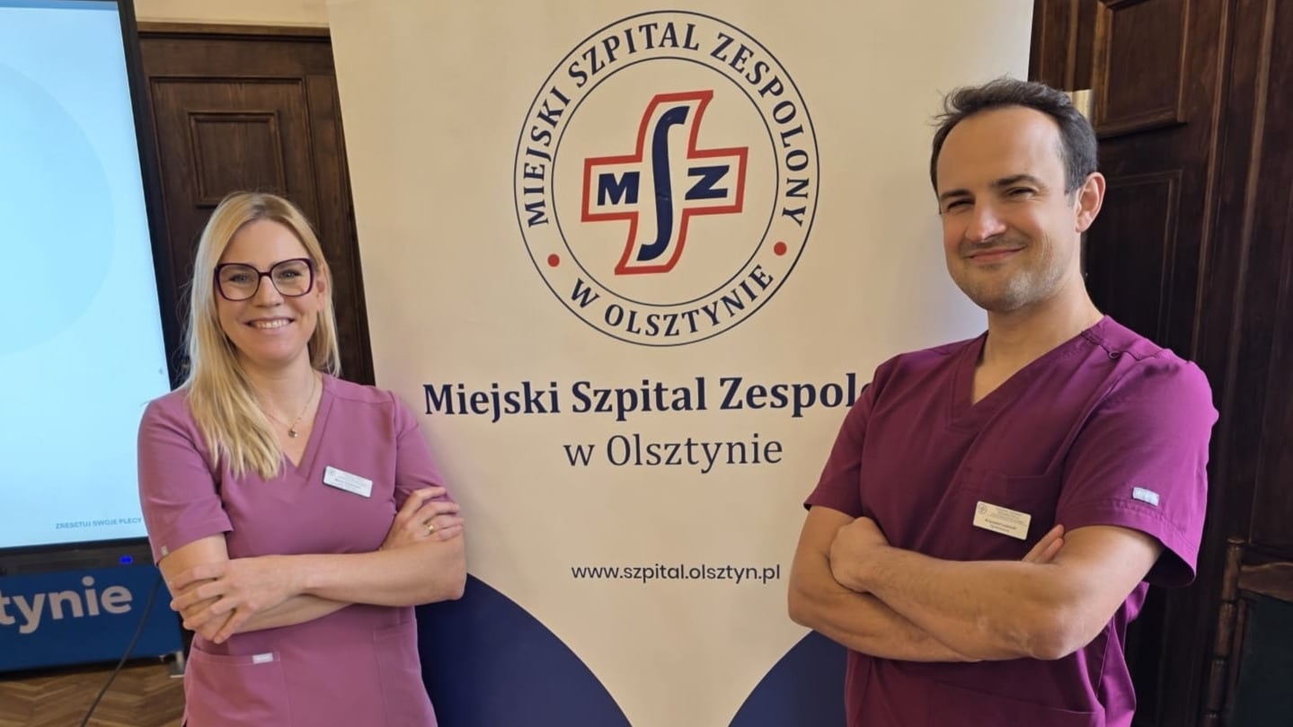 Dwoje pracowników w różowych uniformach stoją obok logo Miejskiego Szpitala Zespolonego w Olsztynie, uśmiechając się.