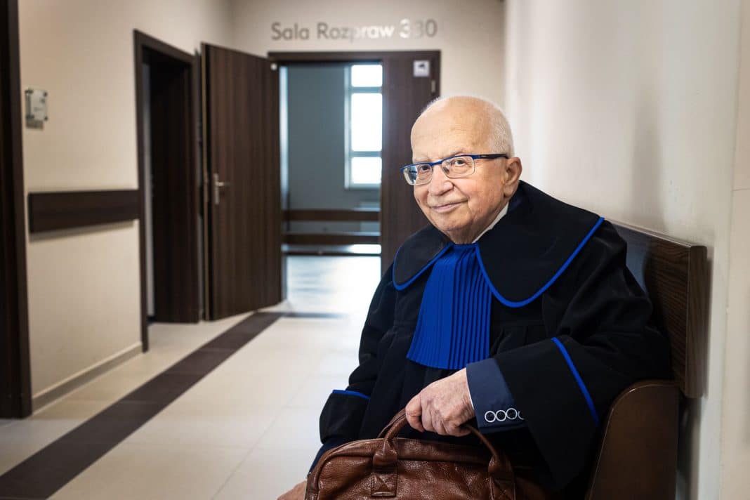 Starszy mężczyzna w togach sędziowskich siedzi na ławce w sądzie, trzymając torbę.