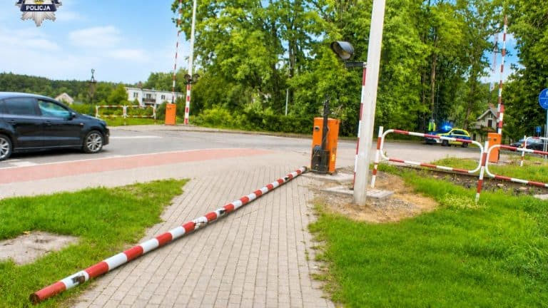 Przejazd kolejowy z opuszczonymi rogatkami, migającymi światłami i nadjeżdżającym pociągiem, widok z perspektywy chodnika.
