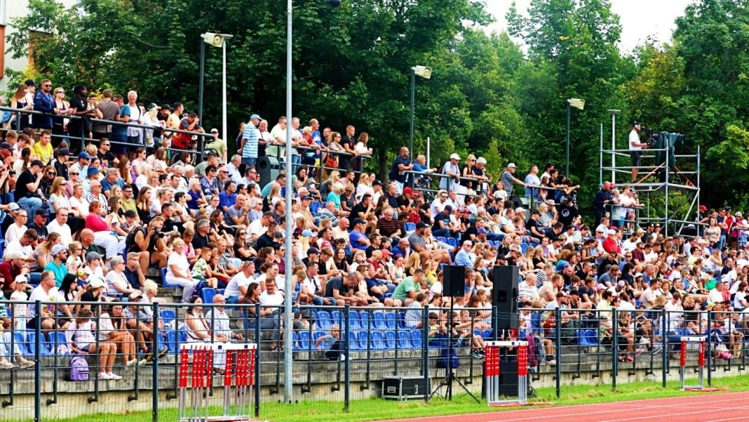 Widok na dużą publiczność siedzącą na trybunach podczas wydarzenia sportowego na stadionie, z zielonymi drzewami w tle.
