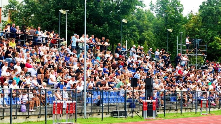 Widok na dużą publiczność siedzącą na trybunach podczas wydarzenia sportowego na stadionie, z zielonymi drzewami w tle.