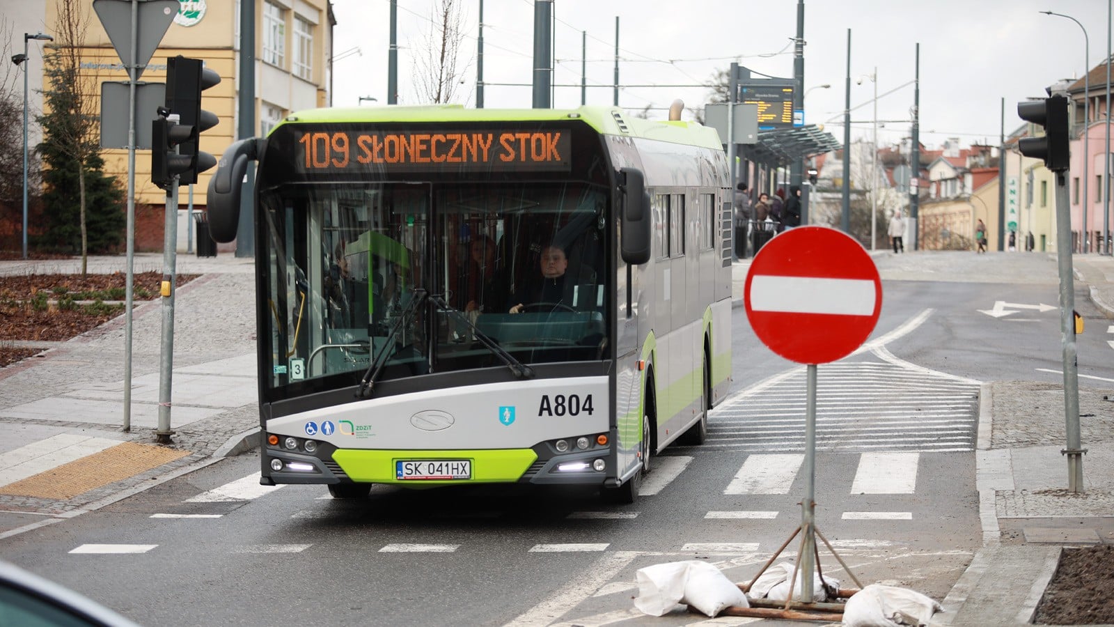 Autobus miejski na przystanku, w tle ludzie czekający na autobus, widoczny rozkład jazdy.