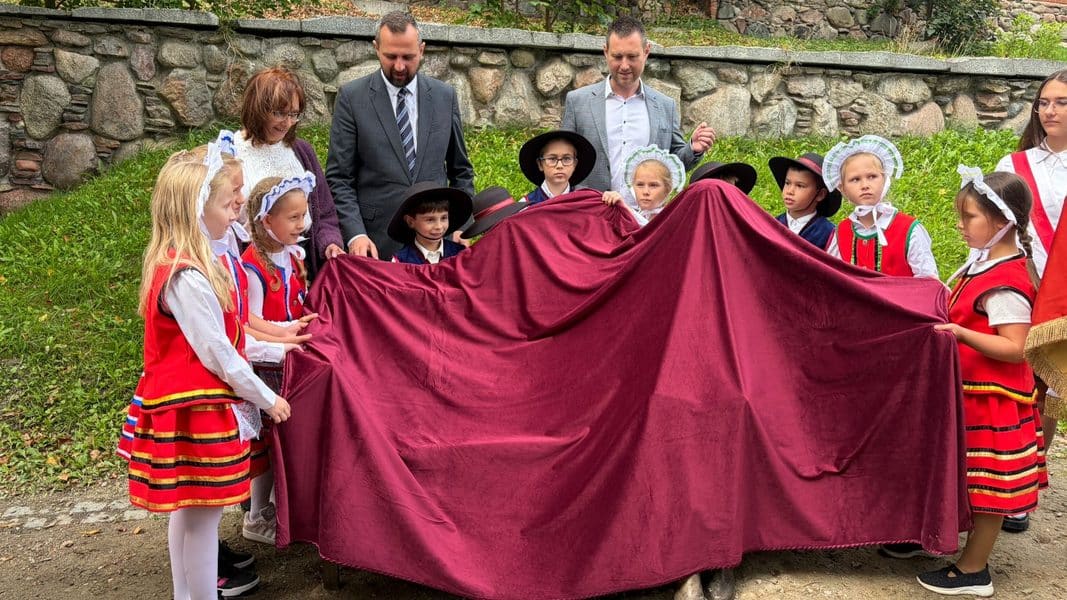 Dzieci w tradycyjnych strojach ludowych podczas odsłonięcia ławeczki w Olsztynie, wokół dorośli.
