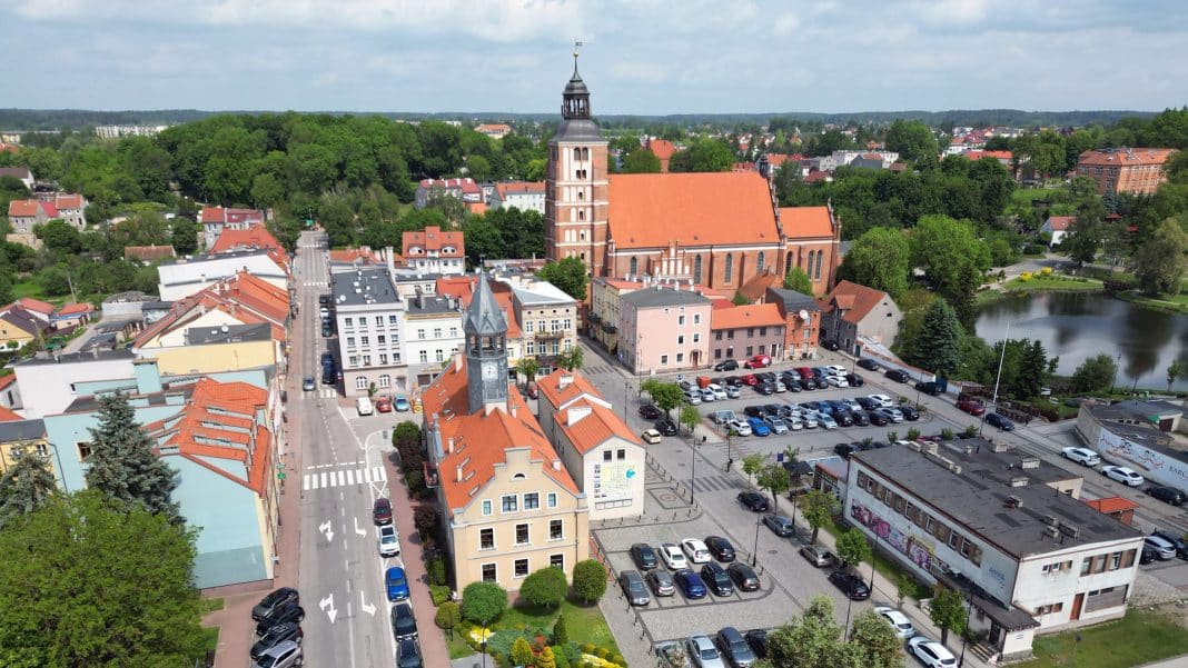 Widok na rynek w mieście z dużym kościołem i parkingiem, otoczony zielenią i budynkami.