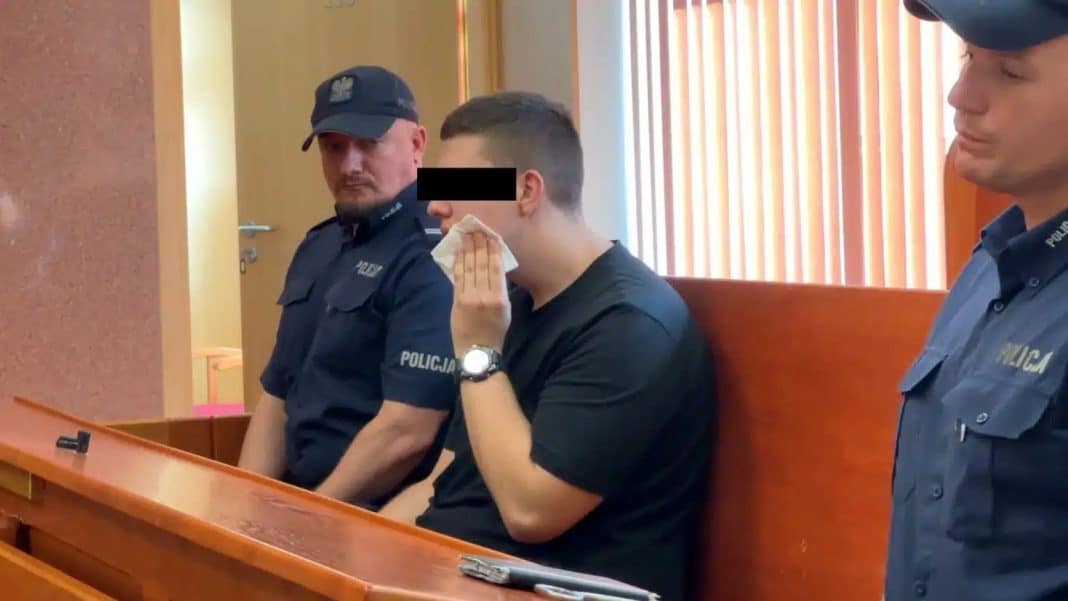 Młody mężczyzna podczas rozprawy sądowej, otoczony przez policjantów.