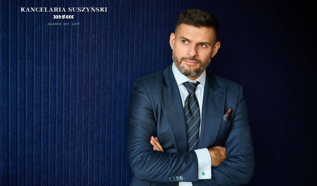Mężczyzna w garniturze stoi z założonymi rękami na tle ciemnoniebieskiej ściany, patrząc w bok.