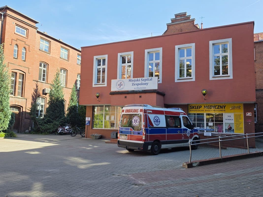 Ambulans stoi przed budynkiem szpitala w Olsztynie, obok sklepu medycznego, na tle czerwonej elewacji.