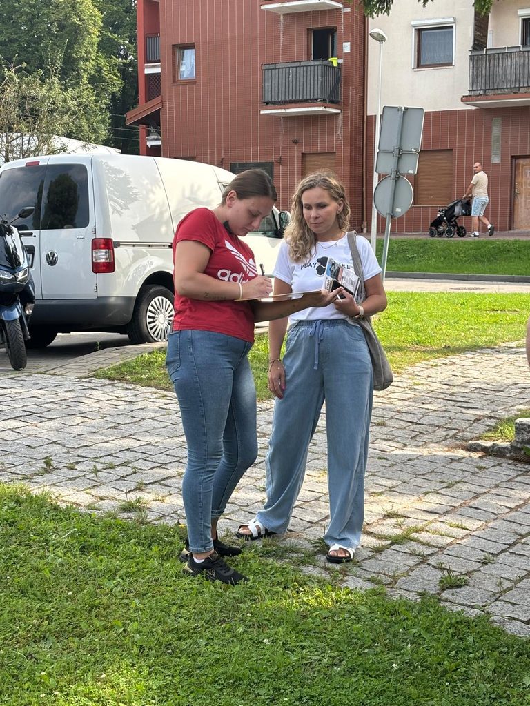 Dwie kobiety zapisują coś na ulicy obok samochodów, w tle budynki mieszkalne i ludzie spacerujący.