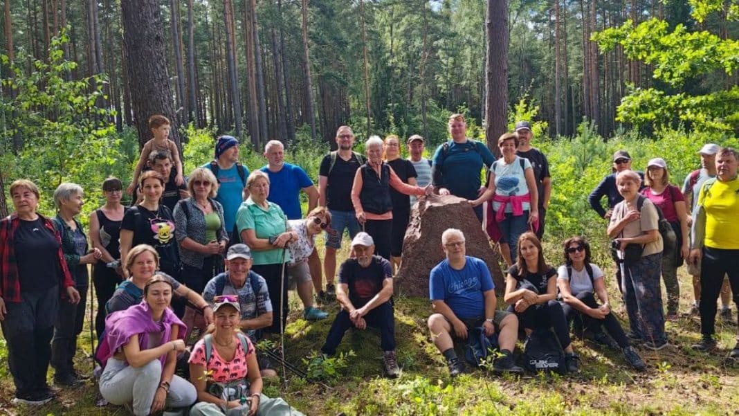 Grupa osób na spacerze w lesie, wokół kamienia, wśród drzew i zieleni.