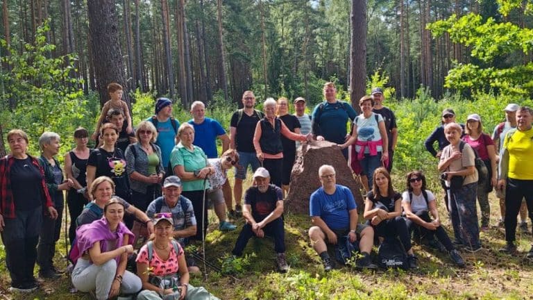 Grupa osób na spacerze w lesie, wokół kamienia, wśród drzew i zieleni.