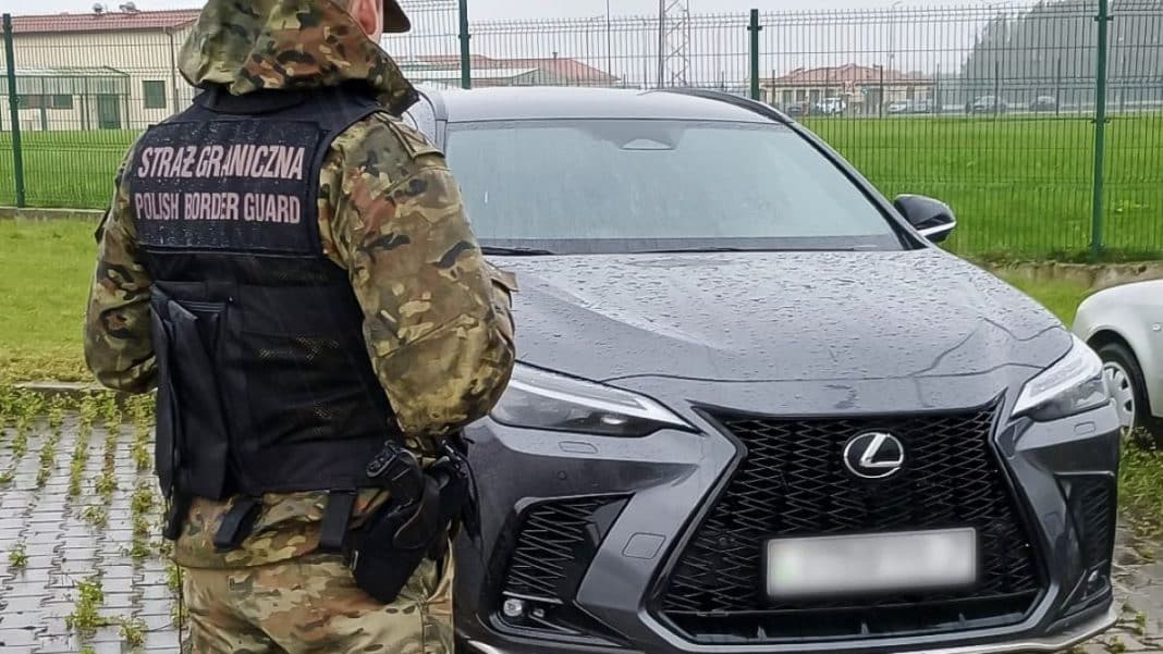 Policjant Straży Granicznej przy luksusowym samochodzie Lexus na granicy, w tle ogrodzenie i budynki.