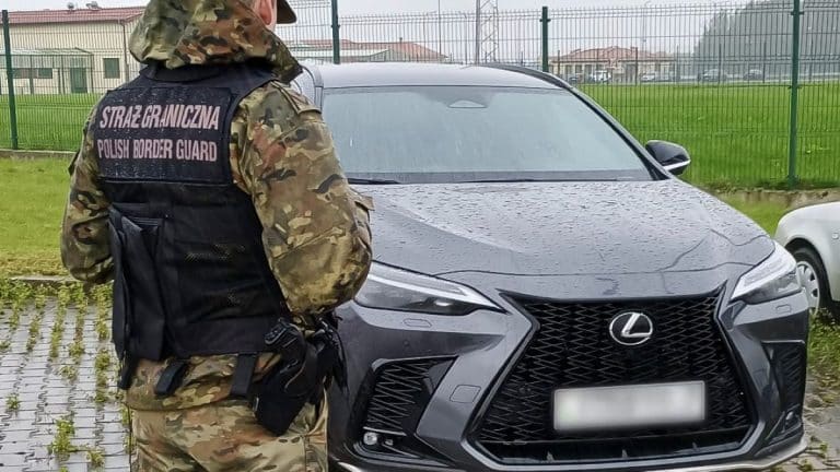 Policjant Straży Granicznej przy luksusowym samochodzie Lexus na granicy, w tle ogrodzenie i budynki.