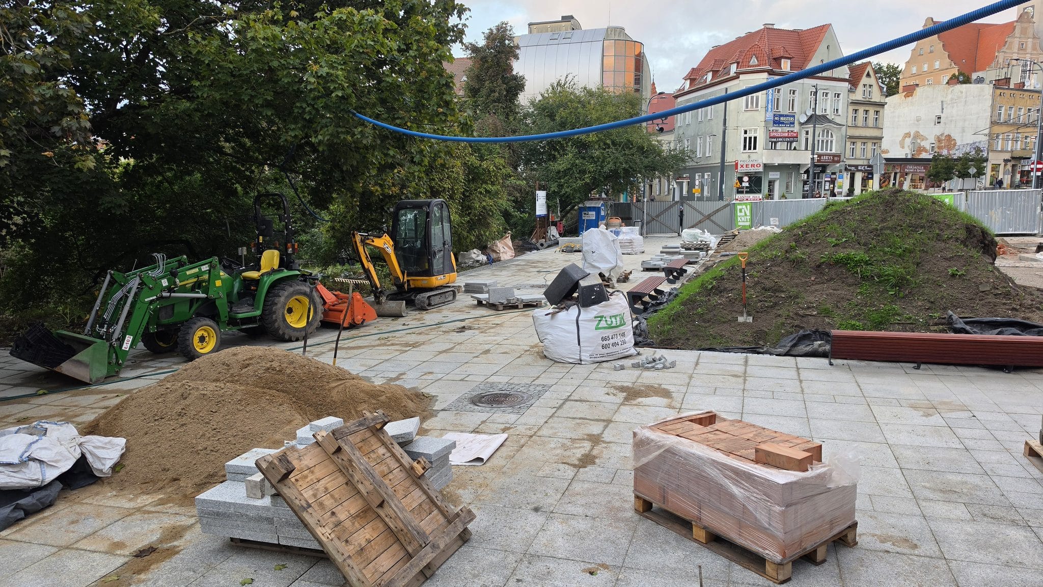 Plac budowy z koparkami, workami z materiałami i kopcem ziemi na tle historycznych budynków.
