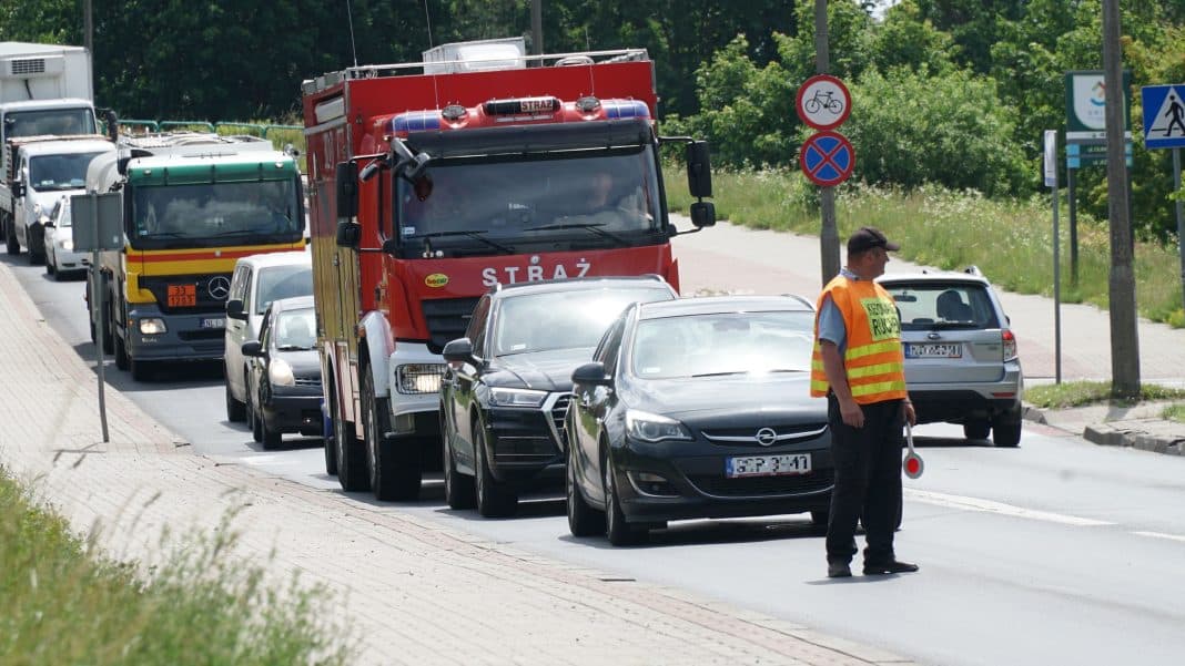 Ciężarówki i samochody stoją w korku na ulicy, obok pracownik z odblaskową kamizelką.