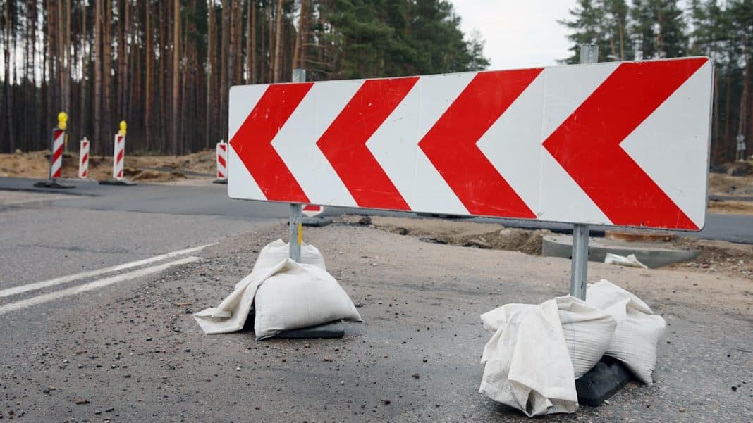 Bariera drogowa z czerwono-białymi pasami na zamkniętej ulicy w trakcie prac remontowych, otoczona workami z piaskiem.