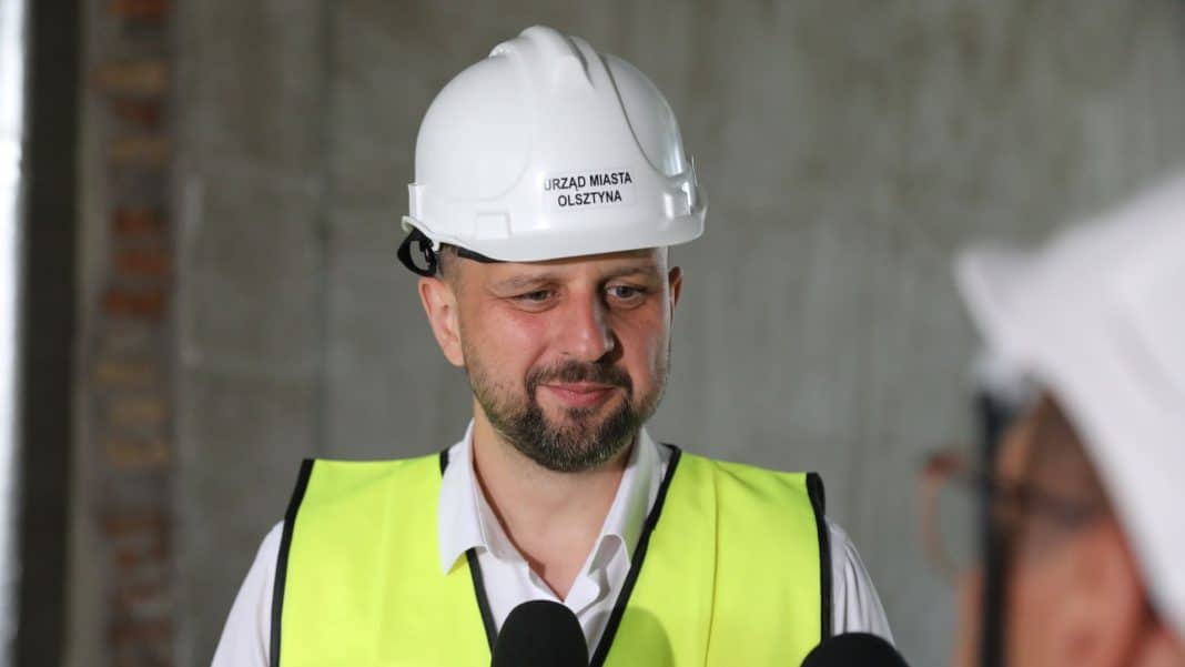 Mężczyzna w kasku i kamizelce odblaskowej podczas wizyty na placu budowy, w tle ściana z cegieł.