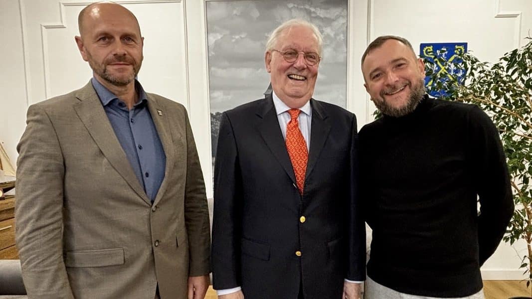 Trzech mężczyzn stojących wewnątrz budynku, uśmiechających się do kamery, w tle obraz i roślina.
