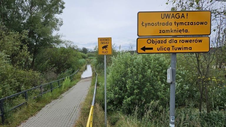 Teren z alejką i tablicami informacyjnymi o zamknięciu ścieżki i objazdach, w tle roślinność i kanał wodny.