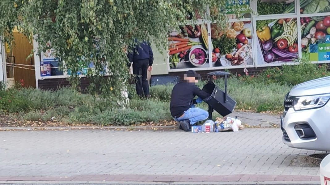Mężczyzna na kolanach przy koszu na śmieci, w tle sklep spożywczy, na chodniku i przy samochodach.
