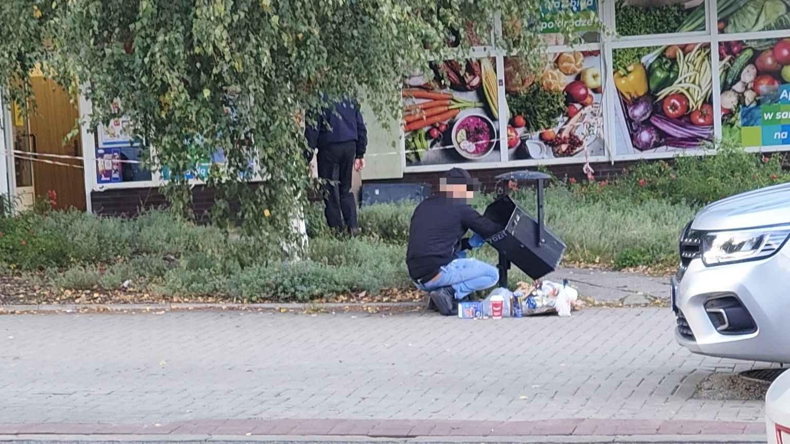 Mężczyzna na kolanach przy koszu na śmieci, w tle sklep spożywczy, na chodniku i przy samochodach.