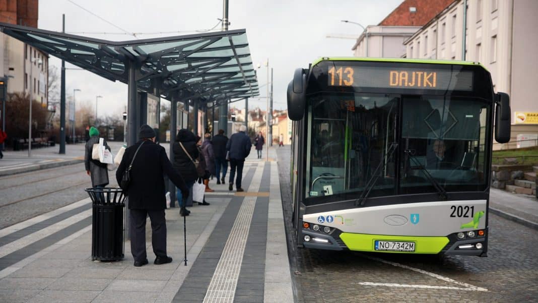 Autobus na przystanku w mieście, ludzie czekają na odjazd, w tle budynki i ulica.