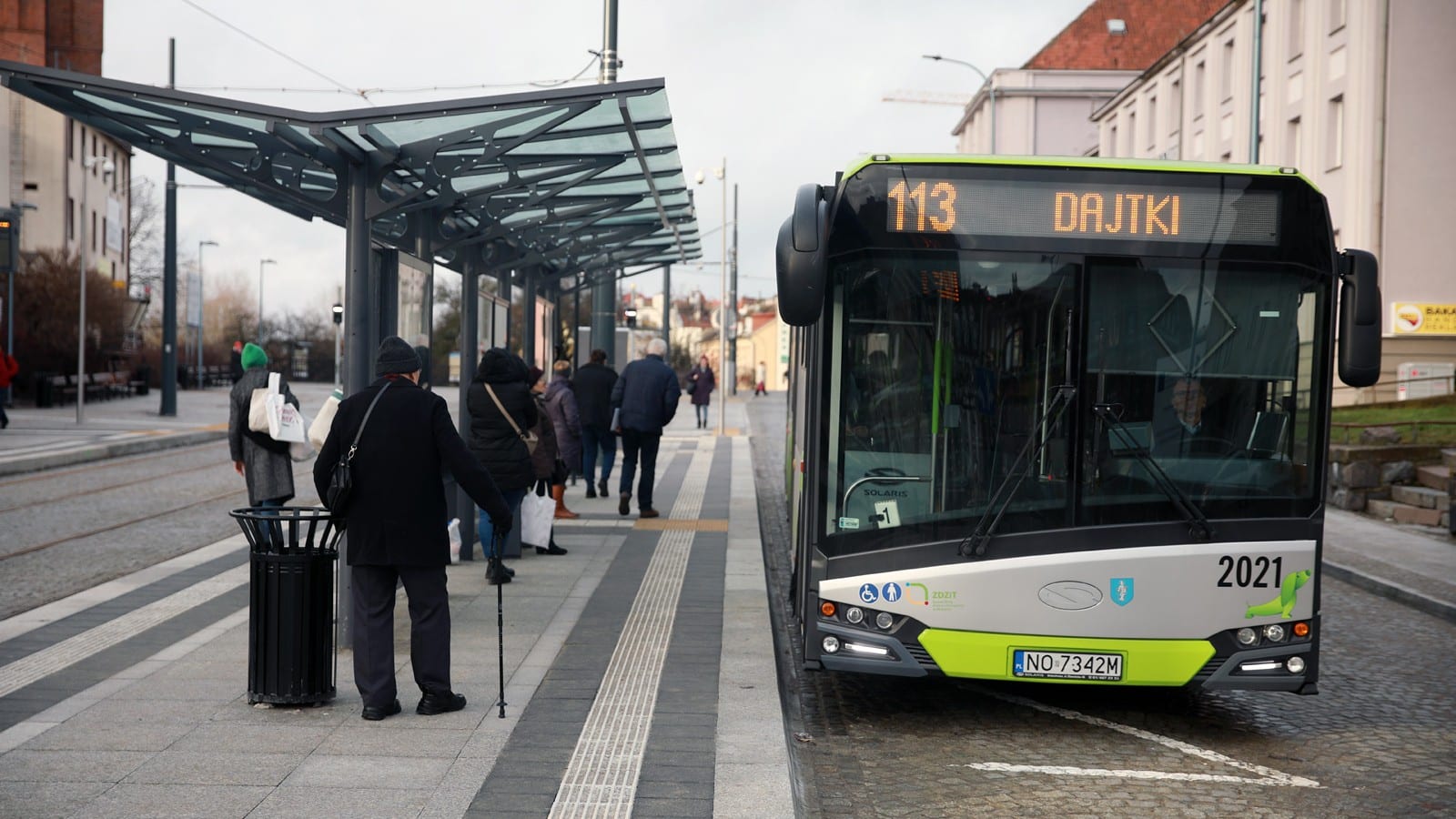 Autobus na przystanku w mieście, ludzie czekają na odjazd, w tle budynki i ulica.