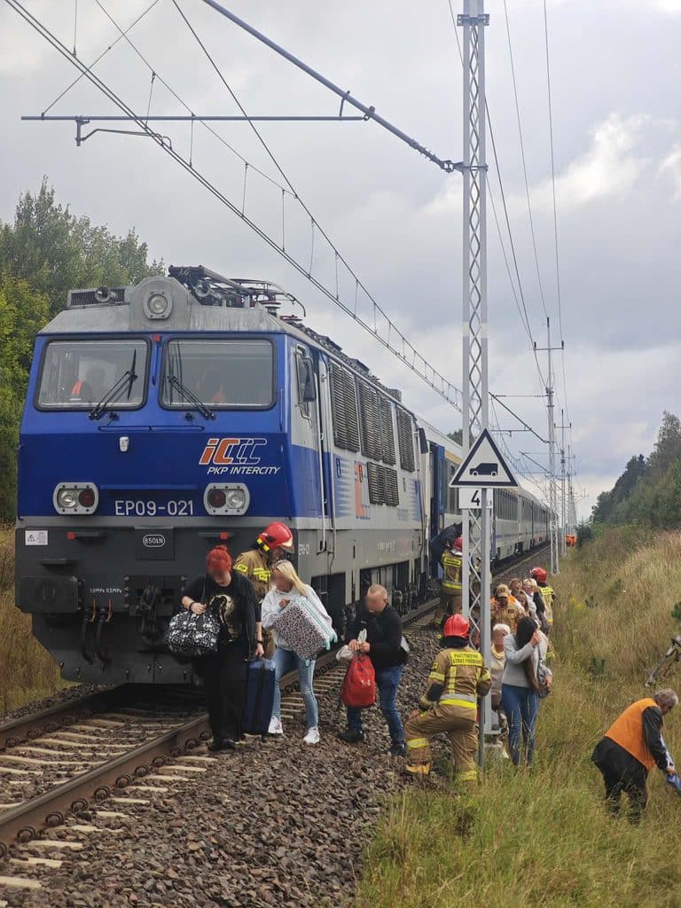 Pociąg PKP Intercity z pasażerami opuszcza tor w trakcie awarii sieci trakcyjnej, wokół strażacy i pasażerowie.
