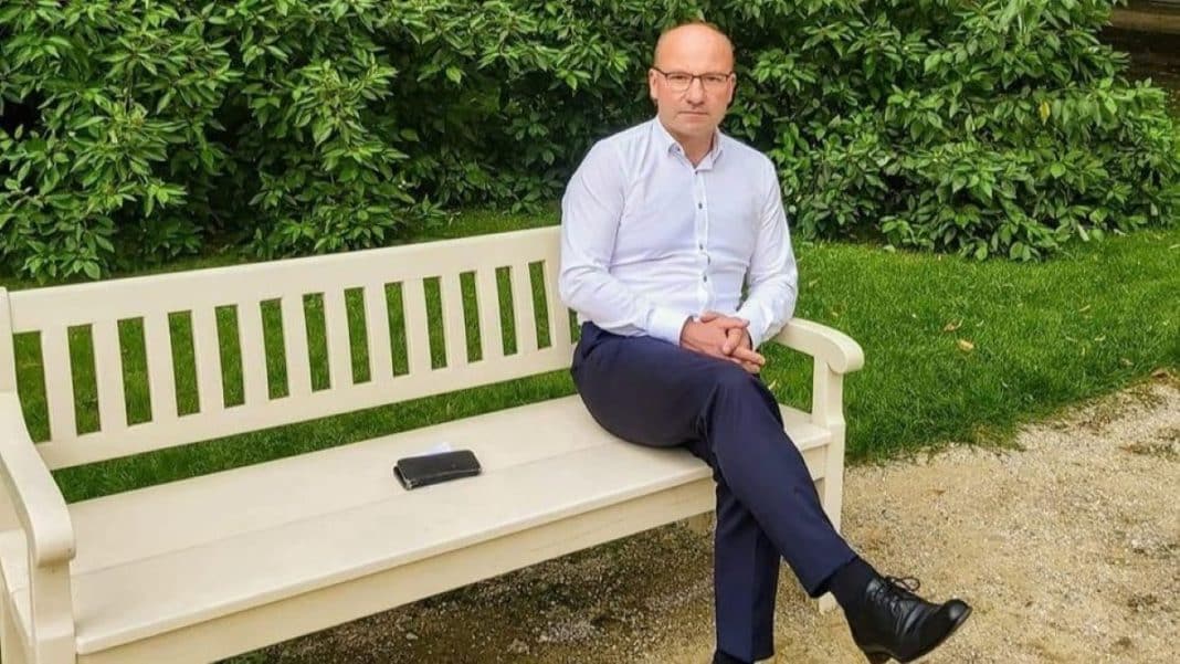 Mężczyzna siedzi na ławce w ogrodzie, ubrany w białą koszulę i ciemne spodnie, obok leży telefon.